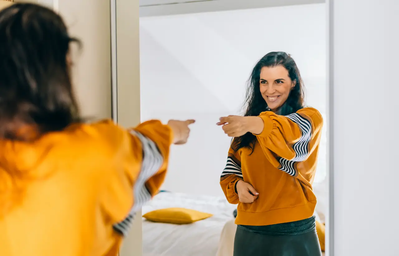 A confident young woman with dark wavy hair wearing a mustard yellow sweatshirt over a black and white striped top, smiling and pointing at her reflection in a bedroom mirror, with a bed and yellow pillow visible in the background.