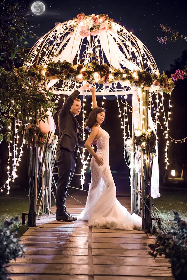 A bride and groom sharing a dance beneath an illuminated ornamental gazebo decorated with pink roses, greenery, and cascading strings of fairy lights. The groom in a dark suit twirls the bride wearing a white lace wedding gown on a wooden walkway, with a full moon visible in the night sky above them.